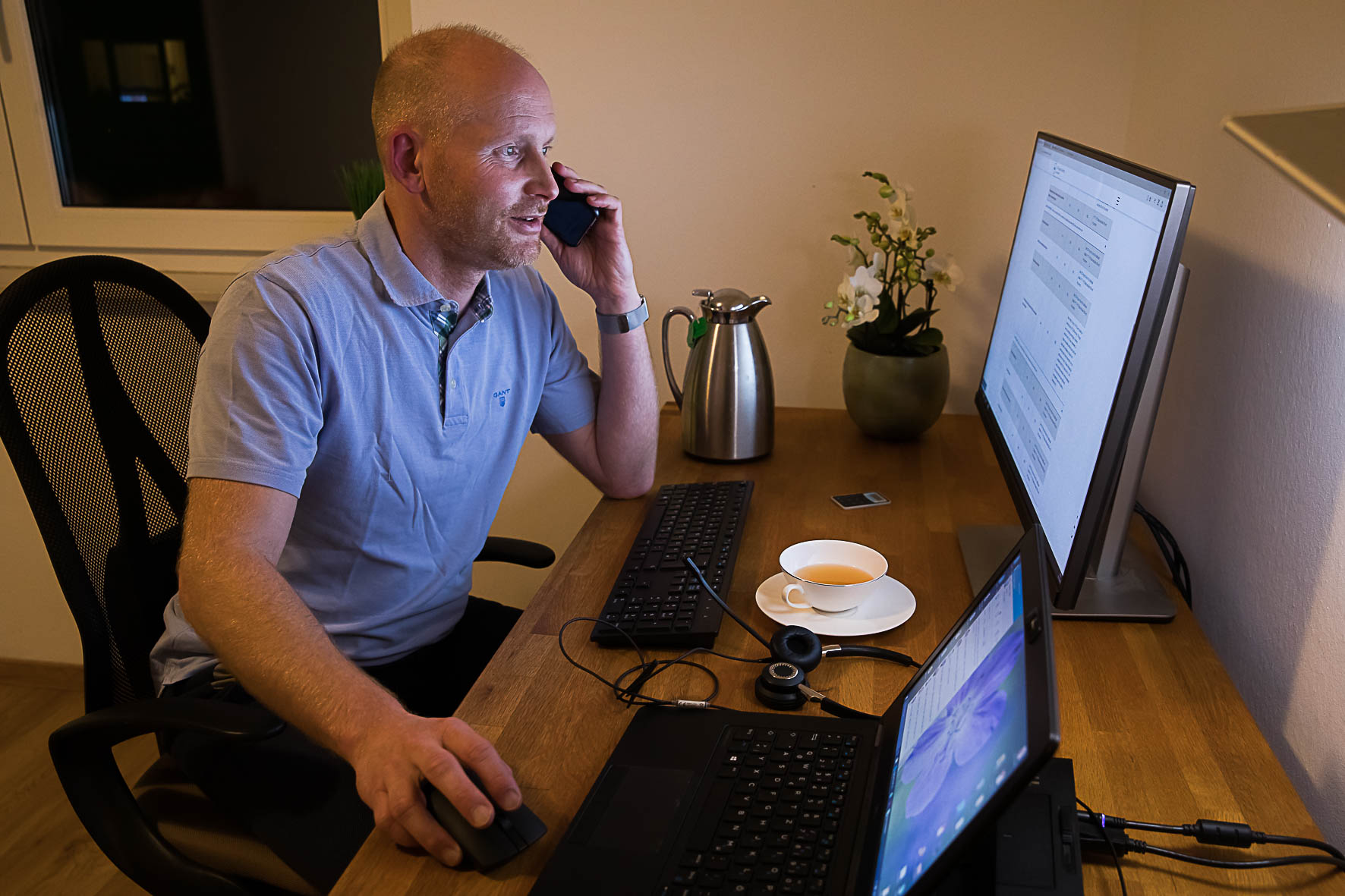 Ein Mann mittleren Alters sitzt mit dem Handy telefonierend an einem Bürotisch mit Bildschirm, Tastatur, Laptop und Headset.