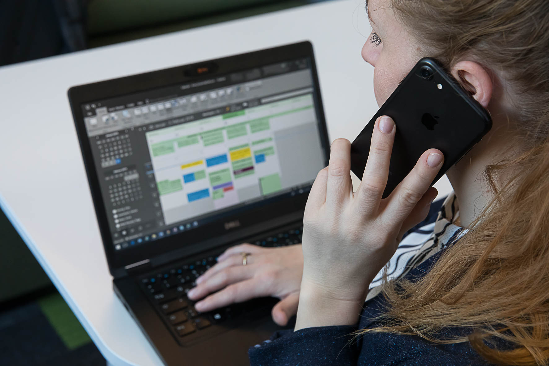 Eine Frau sitzt mit dem Handy telefonierend vor einem Laptop, das einen Kalender mit Wochenplanung anzeigt.