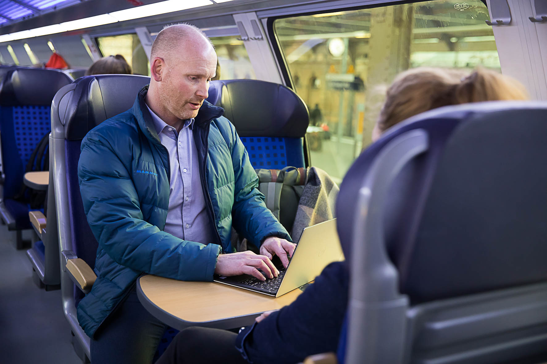 Ein Mann sitzt in der Bahn und arbeitet mit seinem Laptop, das auf einem Tisch steht.