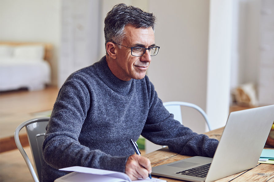 Ein Beschäftigter im Home-Office sitzt am Tisch und arbeitet mit seinem Laptop während er eine Notiz schreibt.