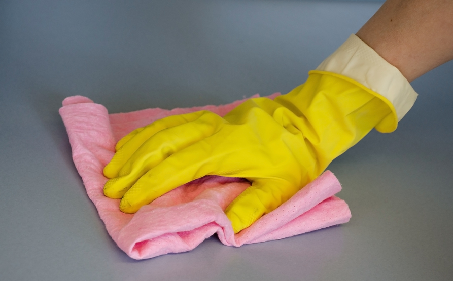 Eine Hand in einem gelben Schutzhandschuh mit umgeschlagener Stulpe wischt mit einem rosafarbenen Lappen über eine graublaue Fläche.